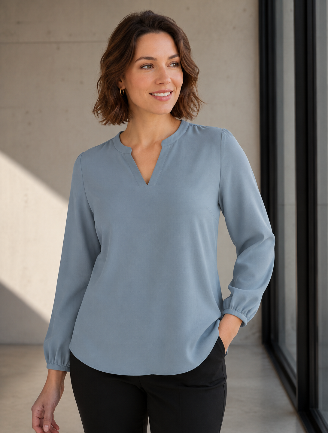 A woman wearing a light blue long-sleeve blouse with a split V-neckline and black pants, standing in a modern indoor setting with concrete walls and large windows.