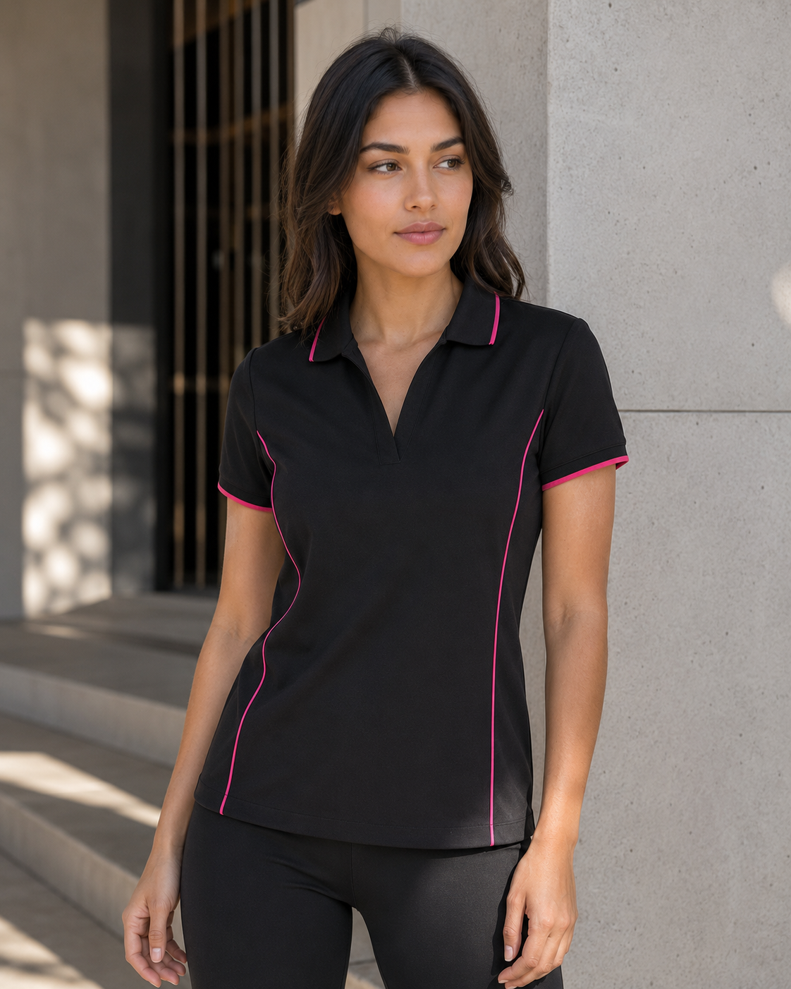 A woman wearing a black short-sleeve polo shirt with bright pink piping details, standing outdoors against a concrete wall.