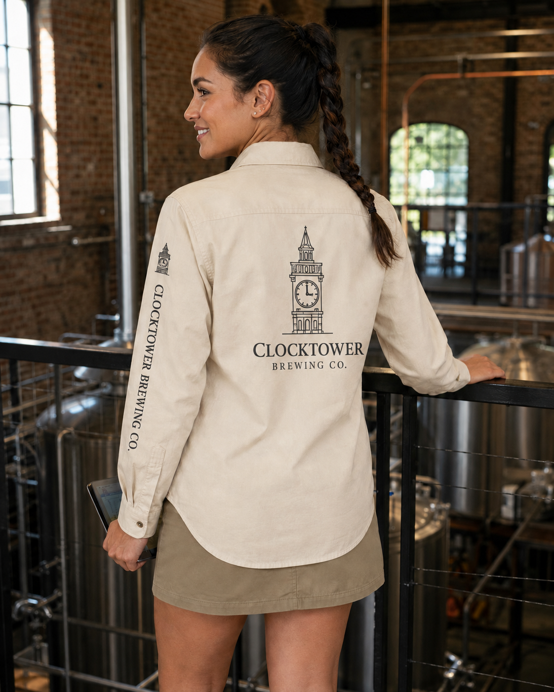 A woman stands in a brewery wearing a cream-colored long-sleeve button-up shirt with a black printed Clocktower Brewing Co. logo on the back and sleeve, paired with a khaki mini skirt.