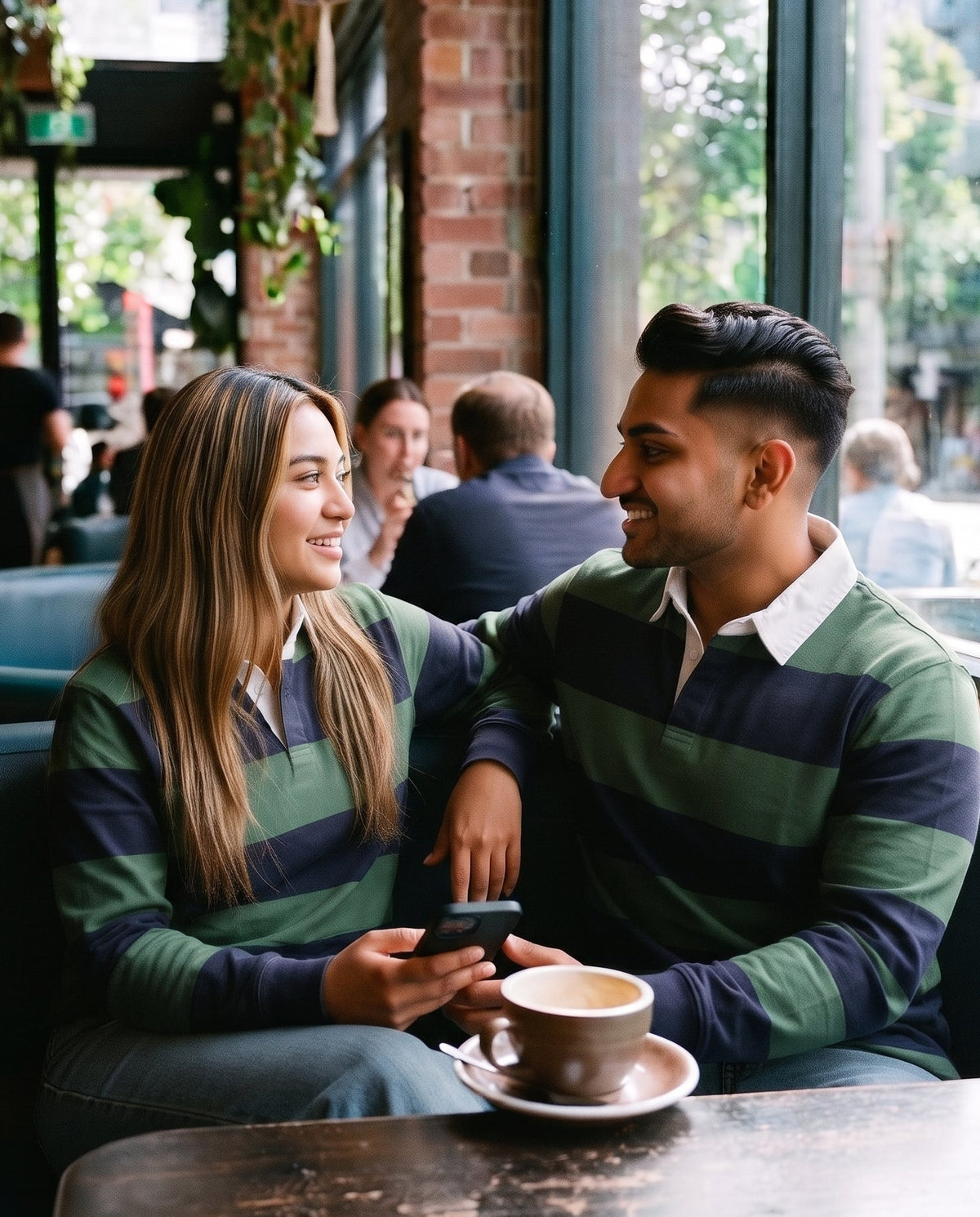 A man and a woman sitting together at a cafe table, both wearing matching green and navy striped long-sleeve shirts. A cup of coffee sits on the table in front of them.