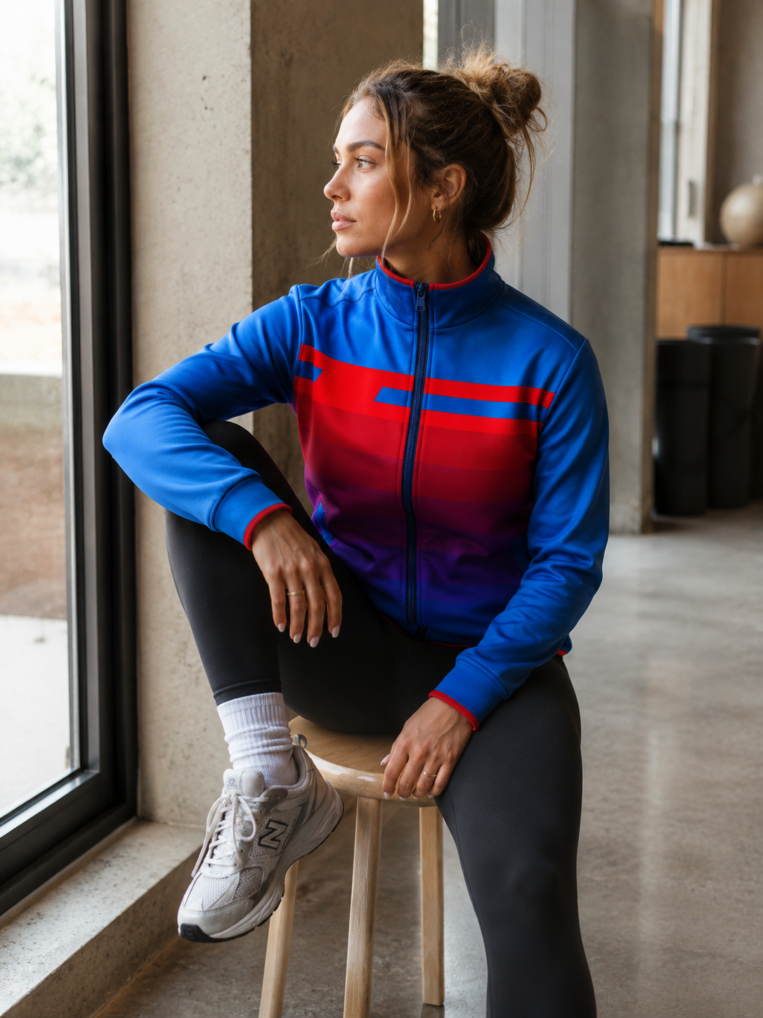 Woman wearing a blue and red jacket sitting on a stool indoors.