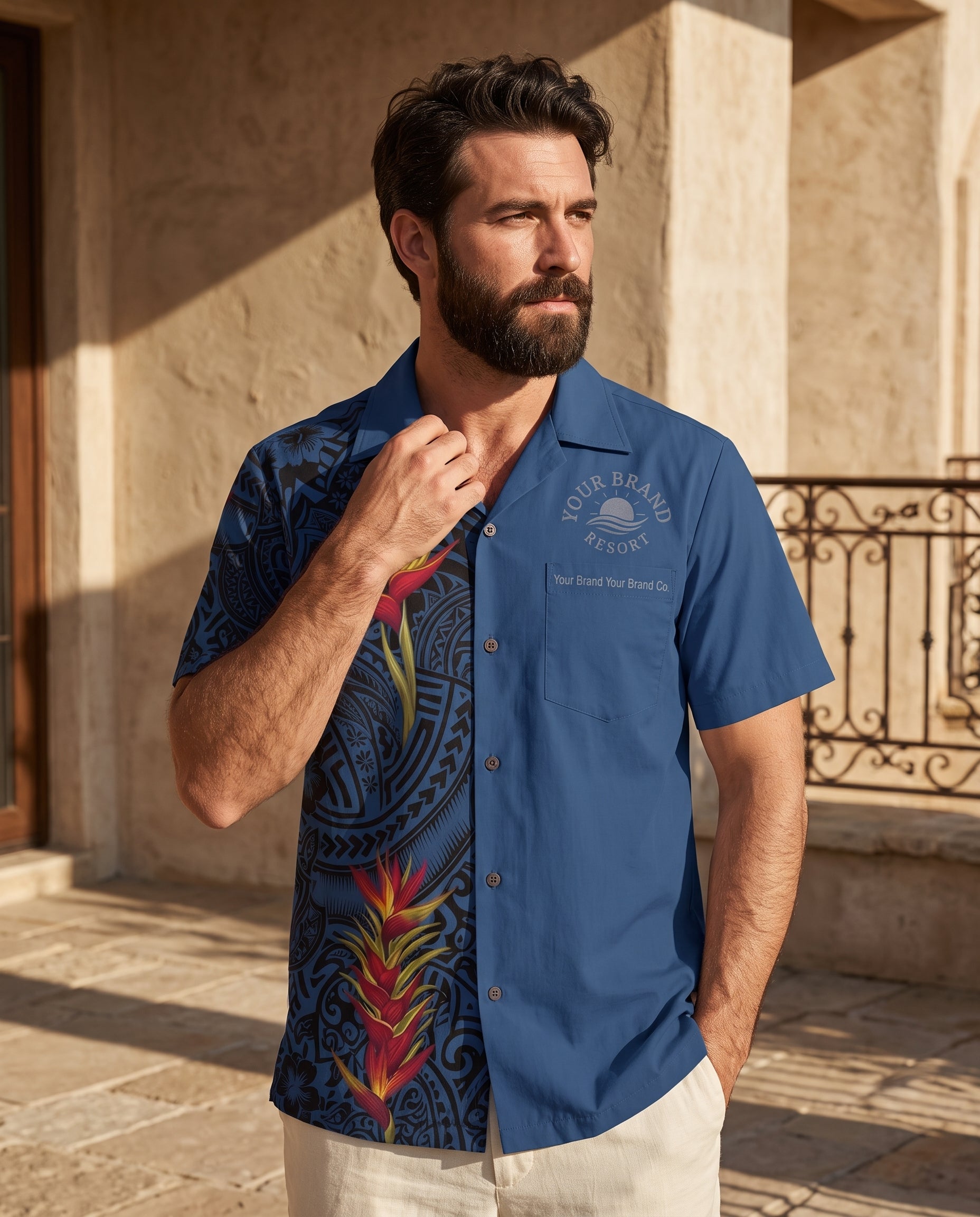 A man wearing a blue short-sleeve button-up camp shirt with a Polynesian tribal print and tropical floral graphics, standing outdoors in front of a beige stone building.