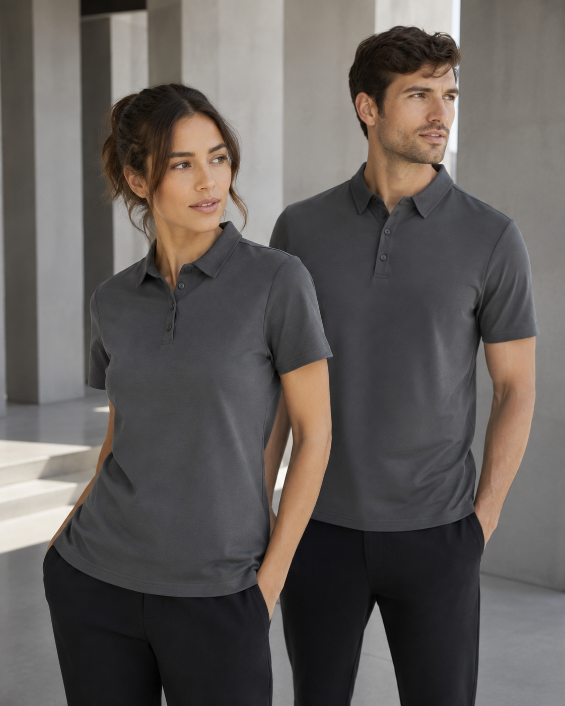 A man and a woman wearing matching dark gray short-sleeve polo shirts, standing in a modern concrete architectural setting.