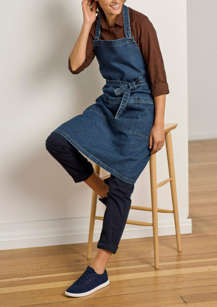 A person wearing a blue denim apron over a brown shirt and dark pants, seated on a wooden stool in a bright indoor room with a light wall and wood floor.