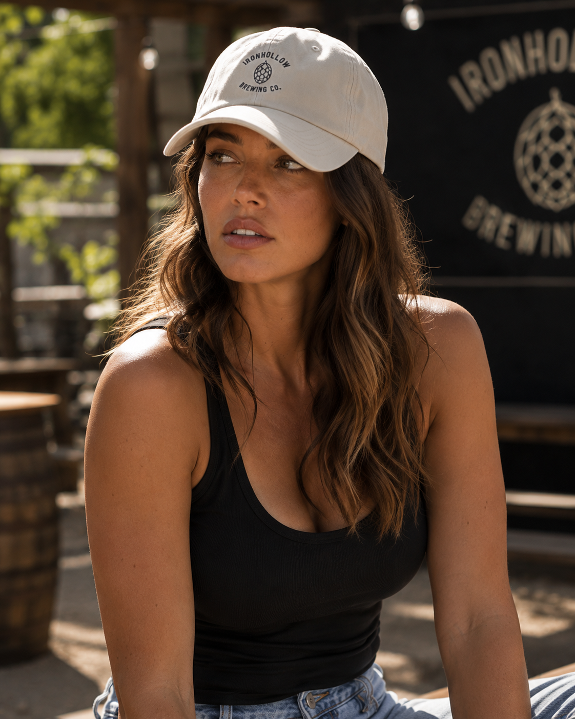 A woman wearing a cream-colored baseball cap with a black circular logo that reads 'Ironhole Brewing Co.' and a black tank top, seated in front of a dark sign with 'Ironhole Brewing' text.