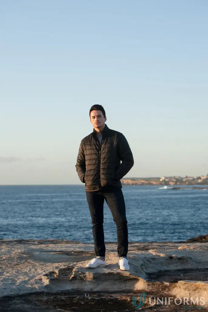 Man in a puffer jacket, dark jeans, and white sneakers on rocky shoreline