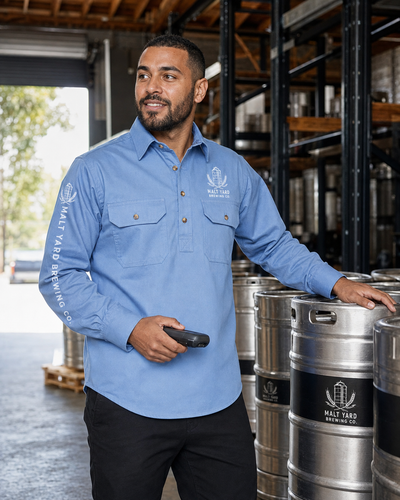 A man wearing a light blue long-sleeve button-up shirt with a printed logo stands in a brewery warehouse next to stainless steel beer barrels.