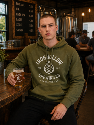 A man wearing an olive green hoodie with 'Ironhollow Brewing Co.' printed in white on the chest, sitting at a wooden bar in a brewery and holding a pint of beer.