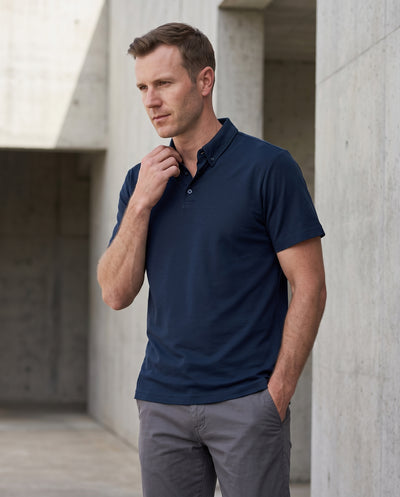 A man wearing a navy blue short-sleeve polo shirt and gray trousers, standing in a modern concrete architectural setting.