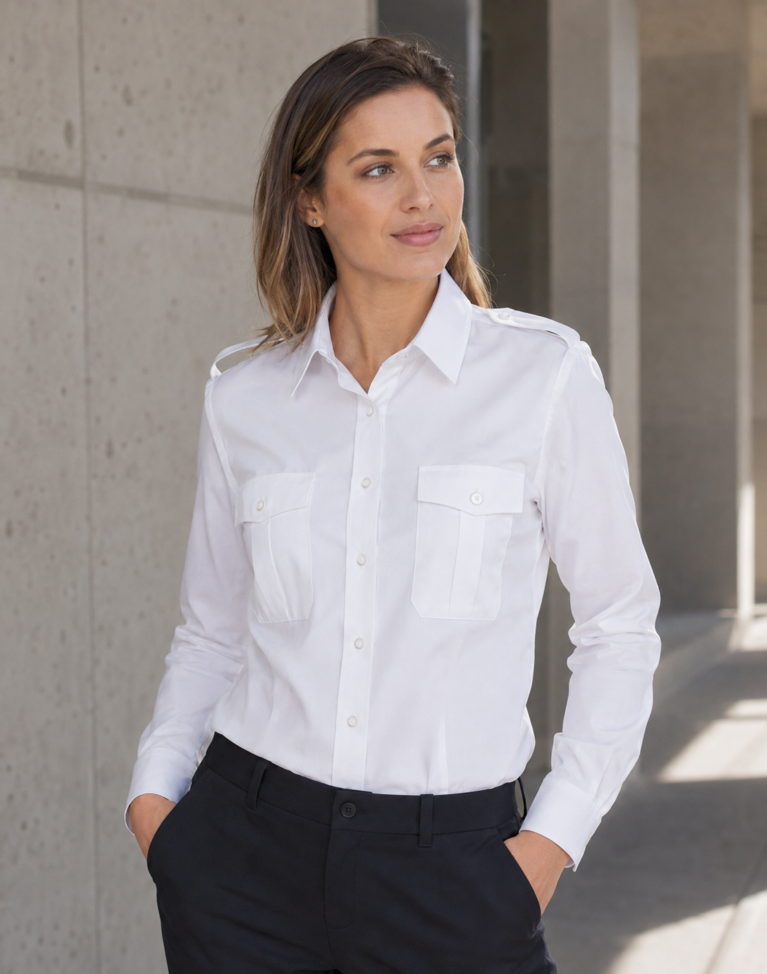 A woman wearing a white long-sleeve button-up shirt with two chest pockets and black trousers, standing in a modern concrete corridor.