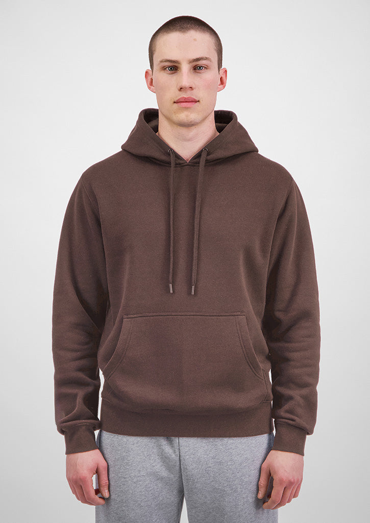 A man wearing a brown pullover hoodie with drawstrings and a kangaroo pocket, paired with light gray sweatpants, standing against a light gray studio background.