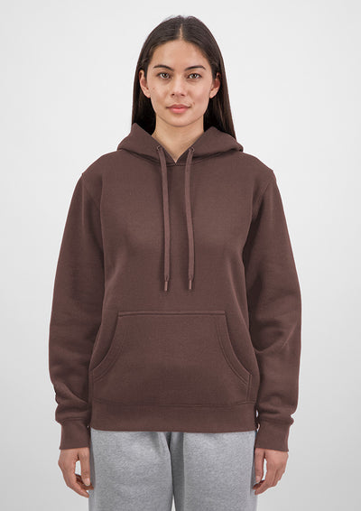 A woman wearing a brown pullover hoodie with matching drawstrings and a front kangaroo pocket, paired with light gray sweatpants, against a plain light gray studio background.