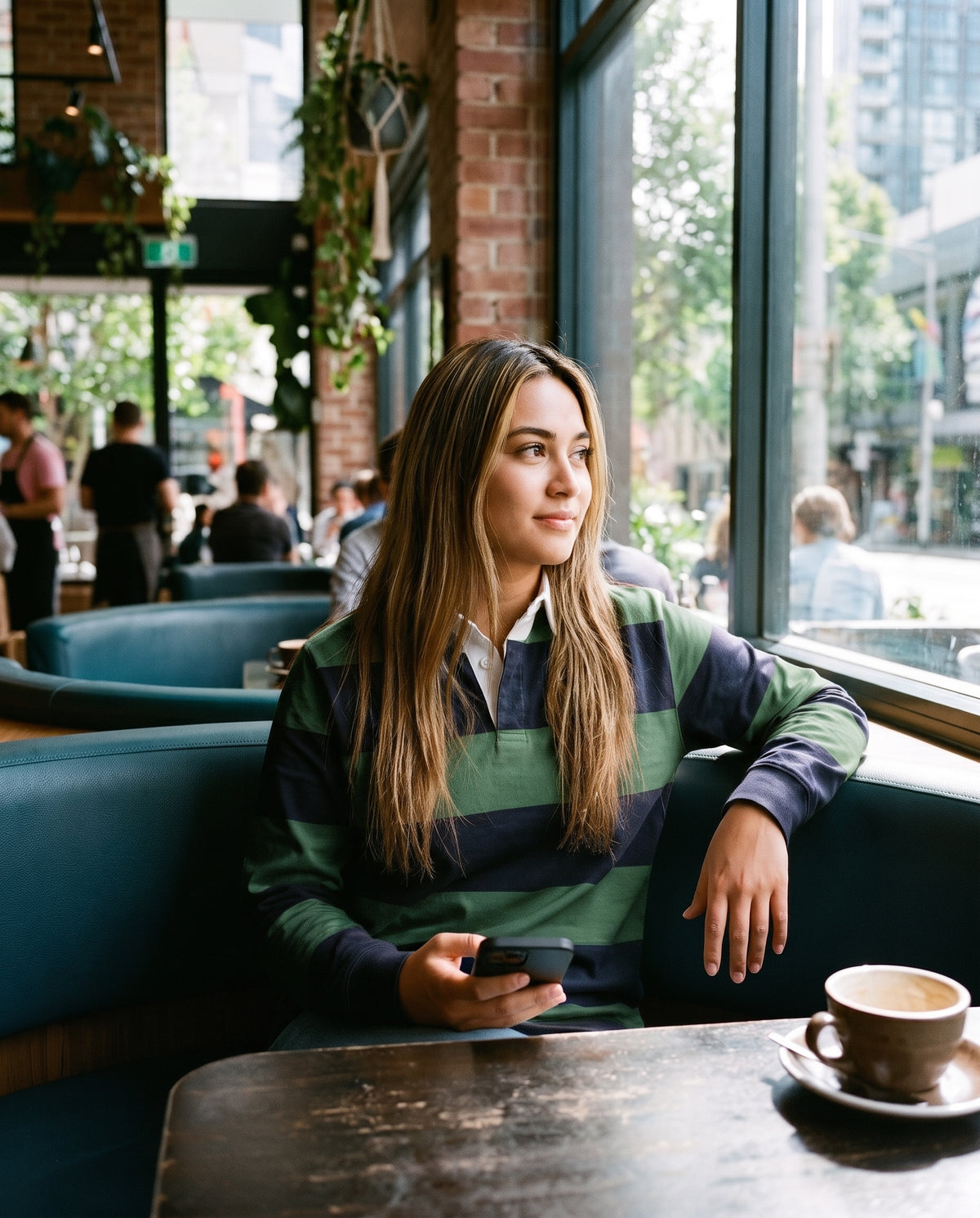 A woman with long light brown hair sits in a teal booth at a cafe, wearing a green and navy striped rugby shirt. She is holding a black smartphone, with a coffee cup on a saucer on the table in the foreground and a brick wall and large windows behind her.