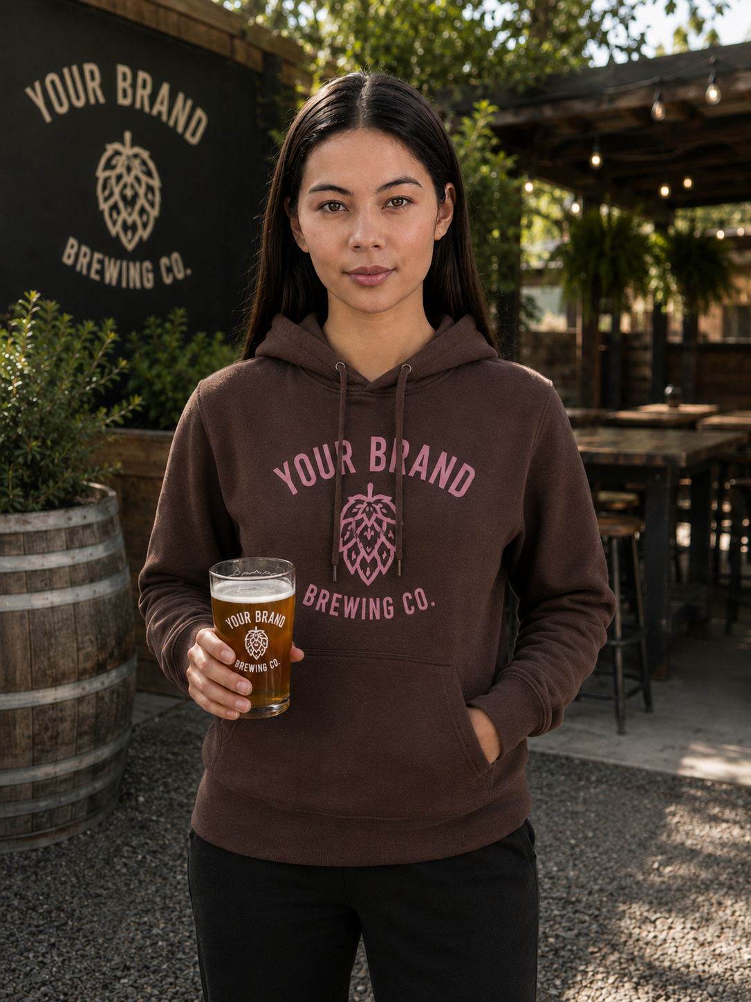 A woman stands outdoors at a brewery holding a pint of beer, wearing a brown hoodie with a pink graphic that reads 'YOUR BRAND BREWING CO.' and a hop cone logo.