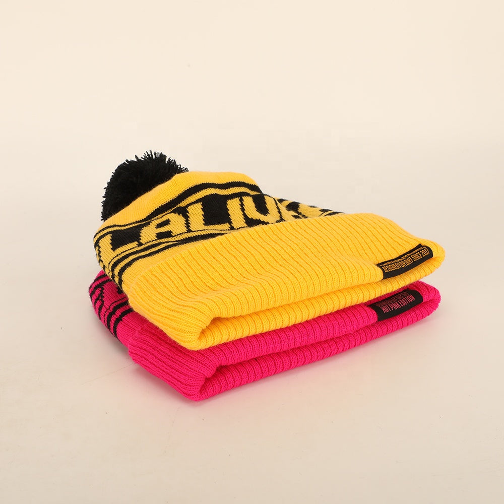 Bright yellow knitted pompom beanie with black lettering stacked on pink knit cap perfect for work uniforms and uniforms