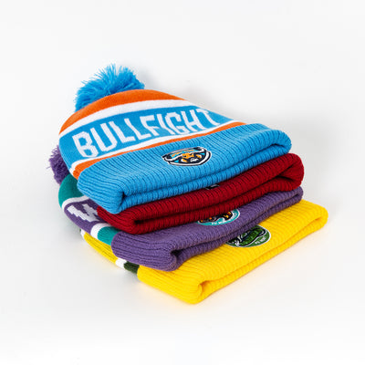 Stack of colorful knitted pompom beanies with blue pompom and bullfight branding perfect for workwear uniforms