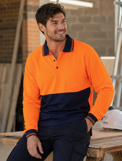 A man wearing a long-sleeve high-visibility polo shirt in bright orange and navy blue, sitting on a wooden workbench in a workshop environment.