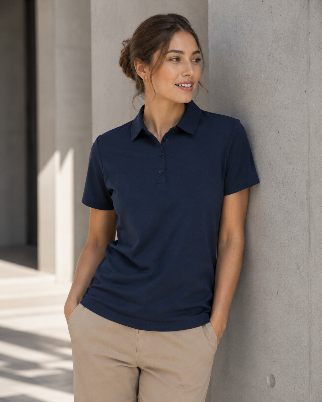 A woman wearing a navy blue short-sleeve polo shirt and khaki pants, leaning against a concrete wall in an outdoor setting.