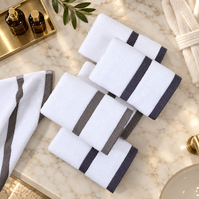 Four folded white towels with dark gray stripes are arranged on a light marble surface, alongside a partially visible matching towel and small bathroom-style props including bottles, a tray, and a bowl.