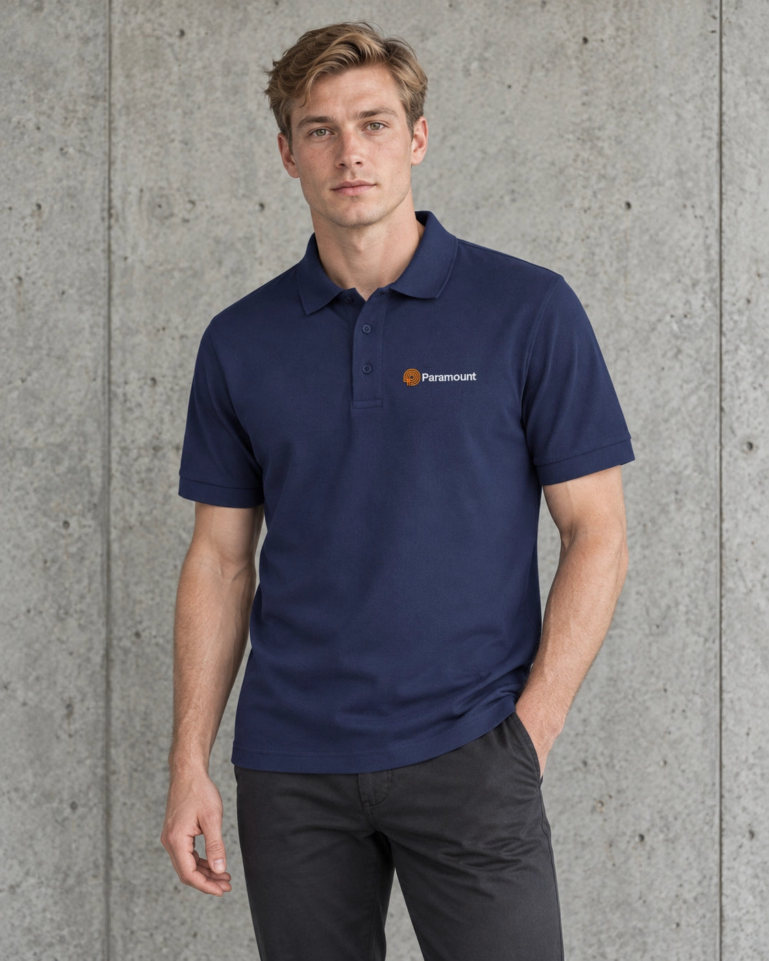 A man wearing a navy blue short-sleeve polo shirt and dark gray pants, standing against a textured concrete wall.