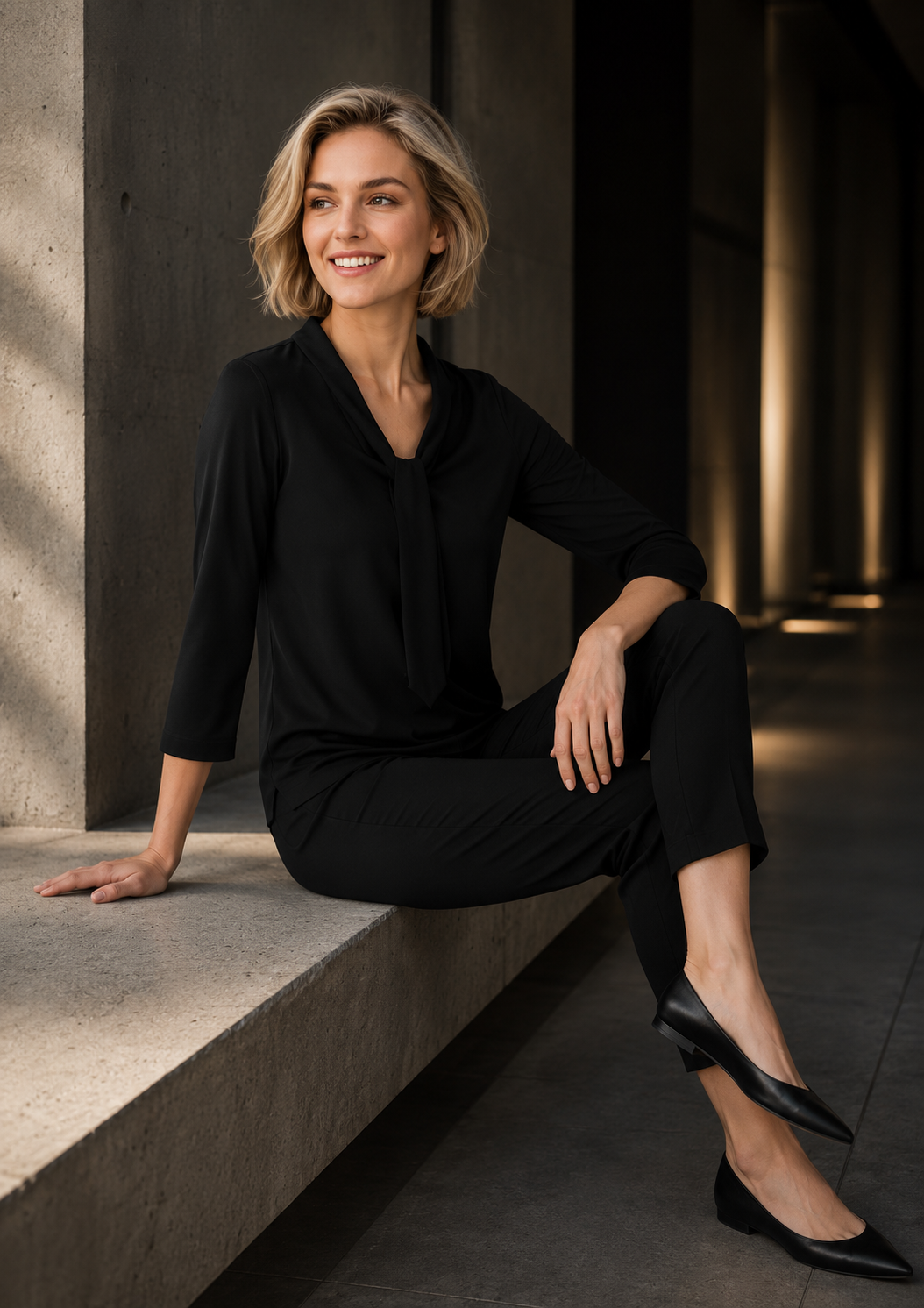 Woman in a black outfit sitting on a concrete ledge with a neutral background