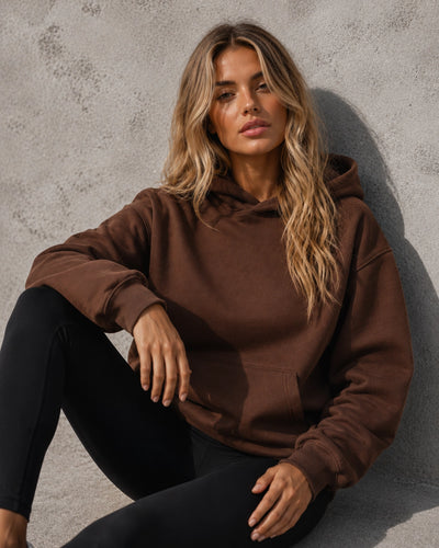 A woman with blonde hair sits against a textured gray concrete wall, wearing a brown oversized pullover hoodie and black leggings.