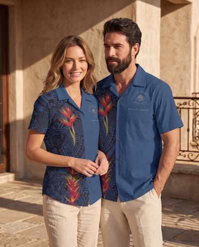A man and a woman wearing matching blue short-sleeve button-up shirts with a dark tropical floral and geometric print, standing outdoors in front of a stone building.