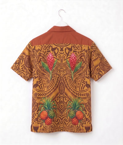 A short-sleeve camp collar shirt shown from the back, hanging on a white hanger against a white wall. The shirt features a warm rust-brown yoke and a back print in a golden-brown tribal pattern decorated with pink tropical flowers and green pineapples.