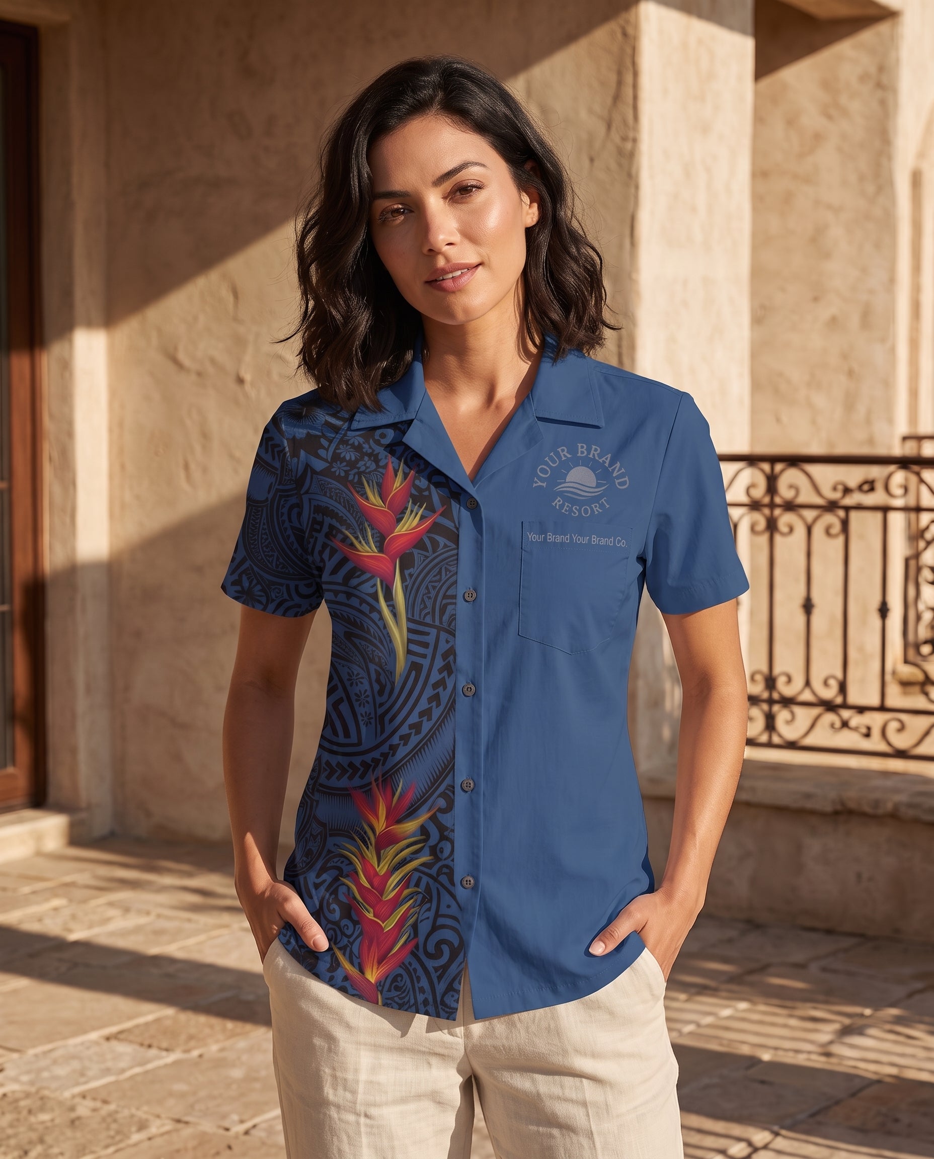A woman stands outdoors wearing a blue short-sleeve button-up shirt with a tropical floral print and a chest logo reading 'YOUR BEYOND HOUSE'. She is also wearing light beige pants, with stone columns and a wrought-iron railing in the background.