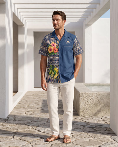 A man wearing a blue short-sleeve button-up shirt with a split design featuring a floral print on one side and a geometric pattern on the other, paired with off-white trousers and sandals, standing in a white architectural walkway.
