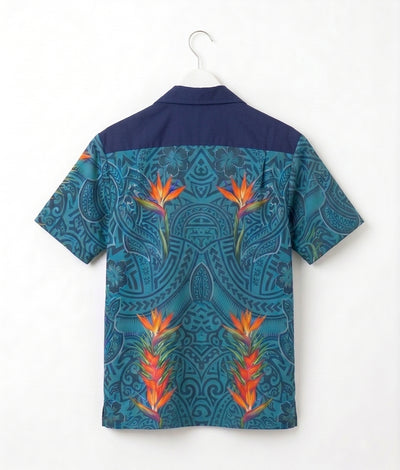 A short-sleeve camp collar shirt shown from the back, hanging on a white hanger against a plain white wall. The shirt features a dark navy blue upper yoke and a lower body with a teal and blue paisley pattern overlaid with three large orange and yellow tropical bird-of-paradise flowers.