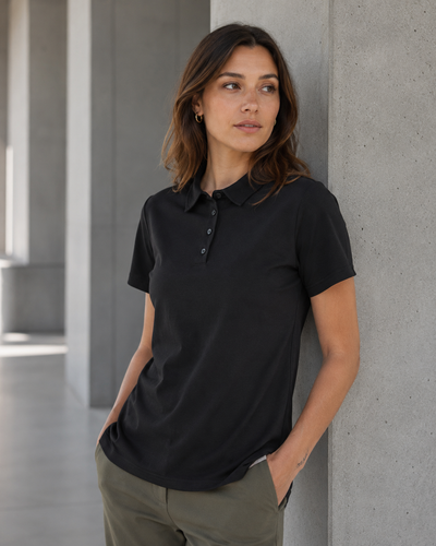 A woman wearing a black short-sleeve polo shirt and olive green pants, leaning against a concrete pillar in a modern architectural setting.