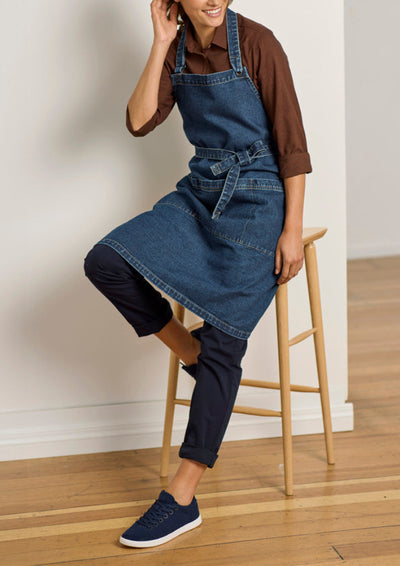 A person wearing a blue denim apron over a brown shirt and dark pants, seated on a wooden stool in a bright indoor room with a light wall and wood floor.