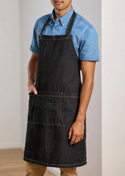 A man wearing a dark charcoal gray denim bib apron over a light blue short-sleeve button-down shirt, standing against a neutral off-white wall.
