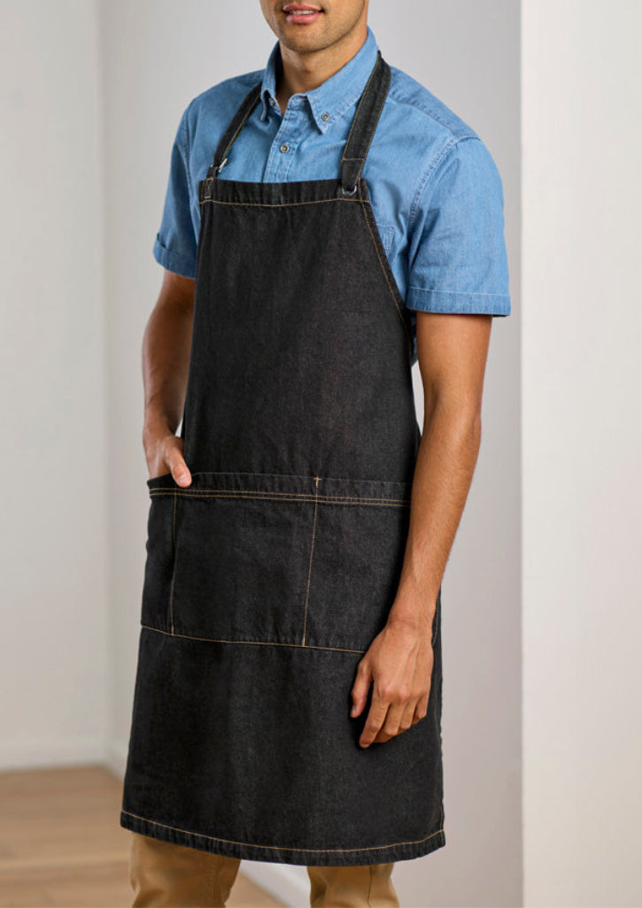 A man wearing a dark charcoal gray denim bib apron over a light blue short-sleeve button-down shirt, standing against a neutral off-white wall.