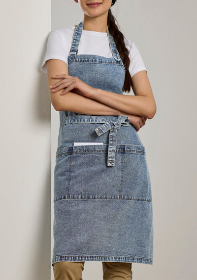 A woman wearing a light blue denim apron over a white t-shirt and tan pants, standing against a neutral beige wall.