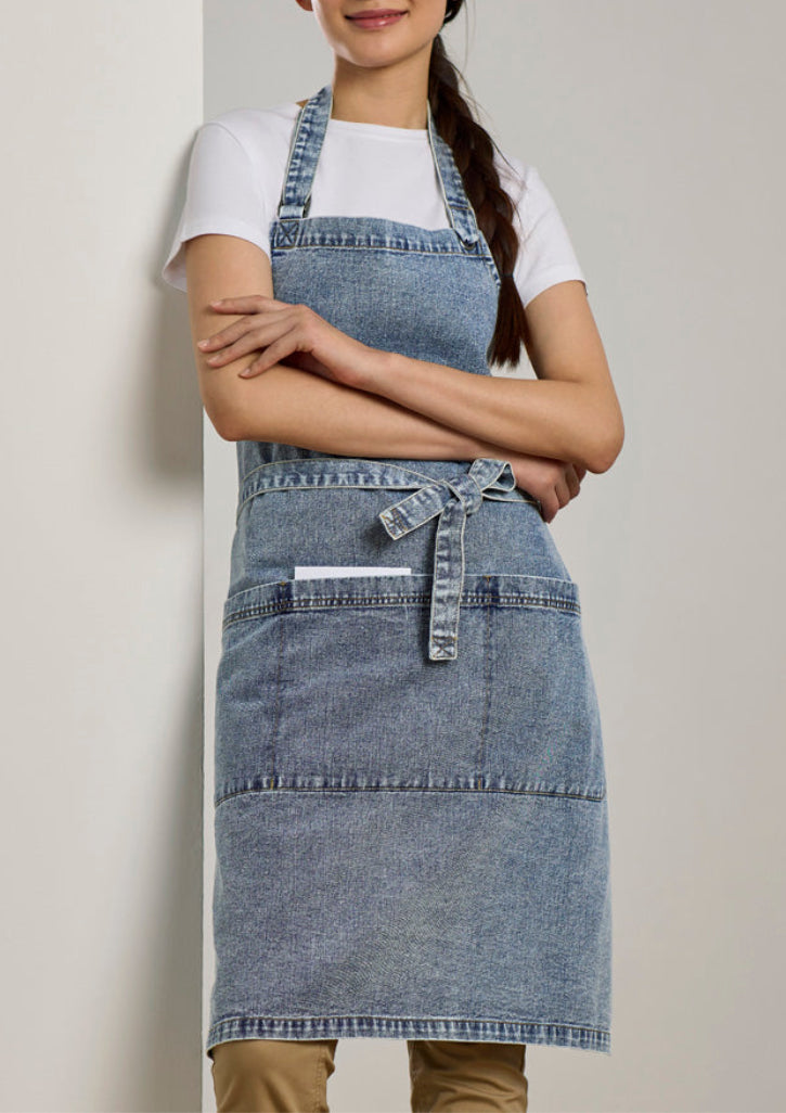 A woman wearing a light blue denim apron over a white t-shirt and tan pants, standing against a neutral beige wall.