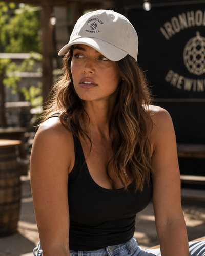 A woman wearing a cream-colored baseball cap with a black circular logo that reads 'Ironhole Brewing Co.' and a black tank top, seated in front of a dark sign with 'Ironhole Brewing' text.