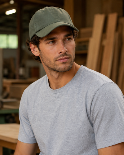 A man wearing an olive green baseball cap and a light gray crewneck t-shirt, standing in a workshop environment.